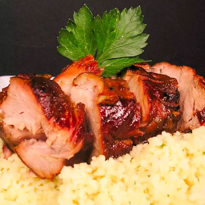 A close-up view of sliced Teriyaki Pork Tenderloin garnished with fresh herbs, served on a bed of couscous.