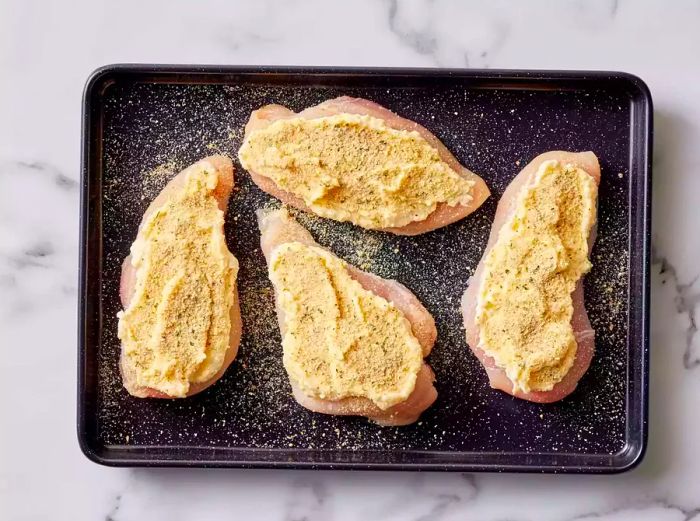 A baking sheet with four chicken breasts topped with a Parmesan-mayo mixture and sprinkled with breadcrumbs