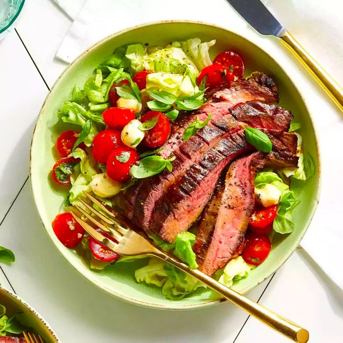 A white bowl filled with Caprese salad, featuring lettuce, tomatoes, mozzarella, and thin slices of grilled flank steak.