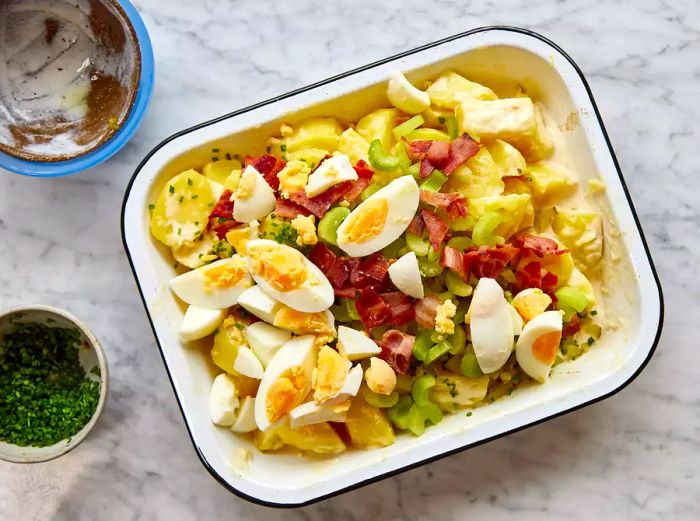 A dish of potato salad featuring celery, chopped pickles, hard-boiled eggs, crispy bacon, and fresh chives