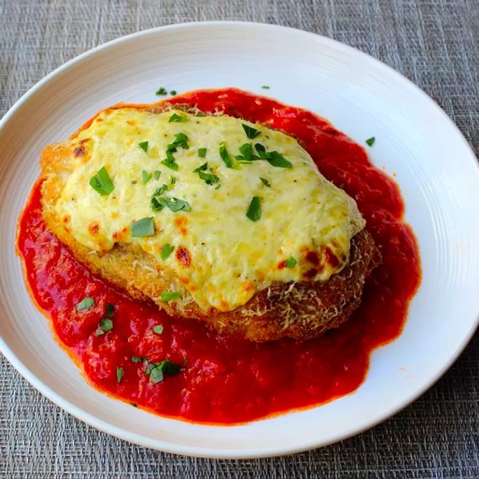 Close-up shot of Chicken Parmesan topped with fresh herbs, served over marinara sauce on a white plate.