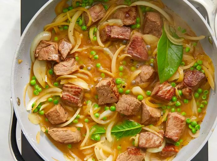 A top-down view of steak, onions, peas, and broth cooking together in a skillet.