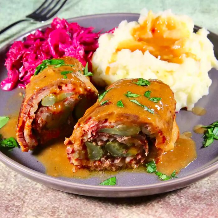 A close-up of Beef Rouladen, topped with savory gravy and fresh herbs, served alongside red cabbage and creamy mashed potatoes with extra gravy on the plate.