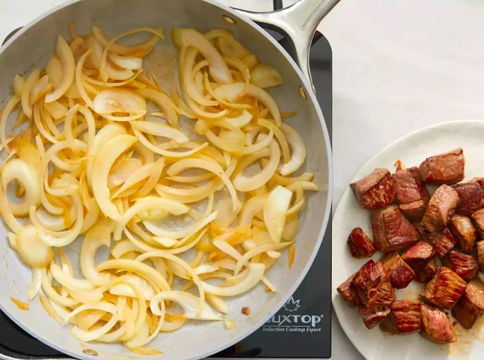 A top-down view of onions sautéing in a skillet, with a plate of cooked steak placed nearby.