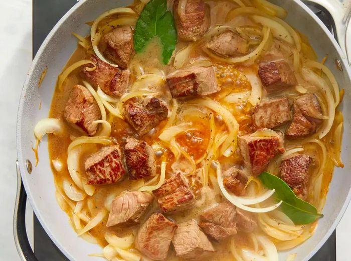 A top-down view of onions, steak, and broth simmering in a skillet.