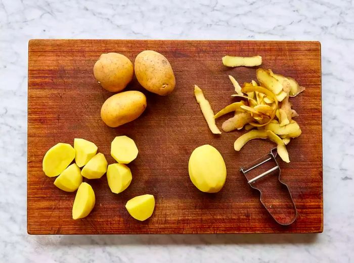 A cutting board with three whole potatoes, a peeled potato, potato skins to the side, and a small pile of sliced potatoes