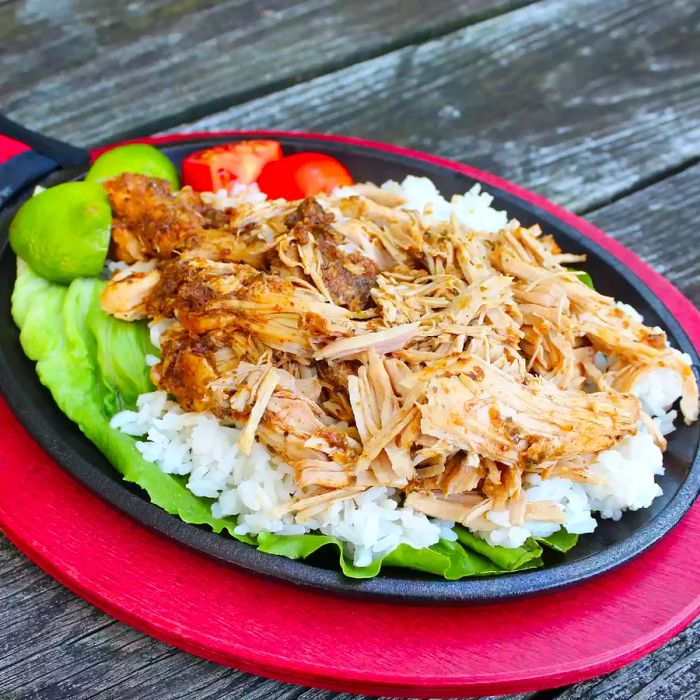 Close-up shot of Slow Cooker Pernil Pork served over white rice and lettuce, topped with lime wedges and tomatoes, in a cast iron skillet.