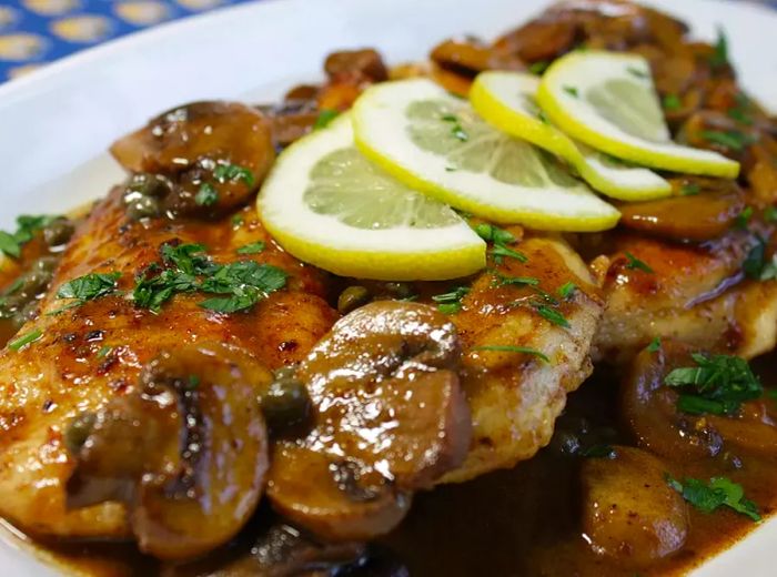 Close-up of a chicken scallopini dish on a white platter, garnished with lemon slices and chopped parsley.