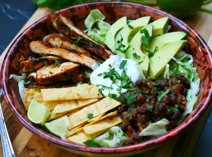 A vibrant salad served in a bowl featuring strips of spiced grilled chicken breast, crispy fried corn tortillas, fresh avocado slices, and a dollop of black bean salsa on a bed of crisp lettuce, topped with sour cream and cilantro for garnish.