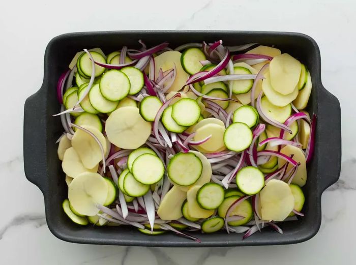 A baking pan filled with chopped red onions, zucchini, and potatoes