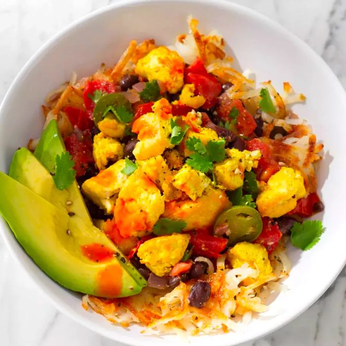 Close-up shot of a Tofu Breakfast Burrito Bowl, garnished with avocado and hot sauce, served in a white bowl.