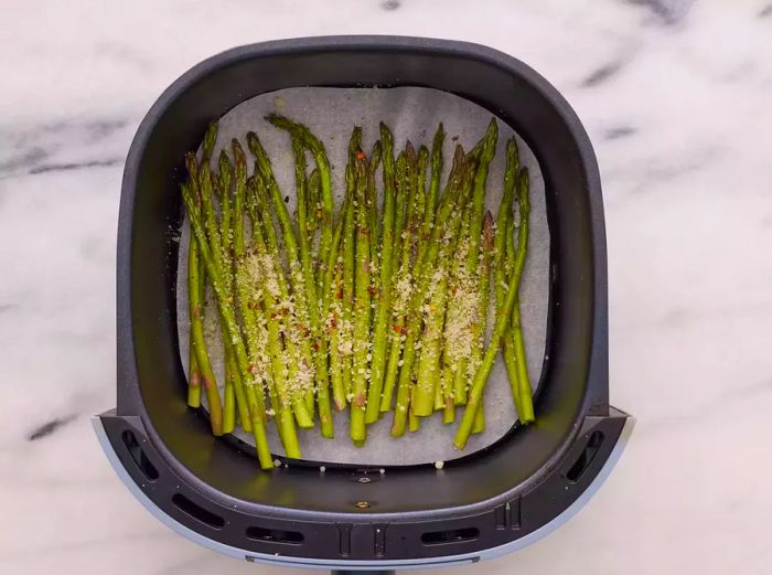 An air fryer basket lined with parchment paper, filled with asparagus and topped with garlic, salt, pepper, chili flakes, and Parmesan cheese.