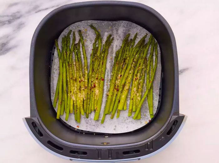 An air fryer basket lined with parchment paper, filled with roasted asparagus and seasoned with garlic, salt, pepper, red chili flakes, and Parmesan cheese.