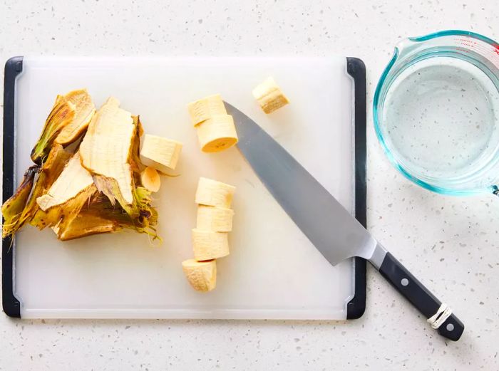 A cutting board with a peeled plantain, sliced into rounds, and a measuring cup filled with water.