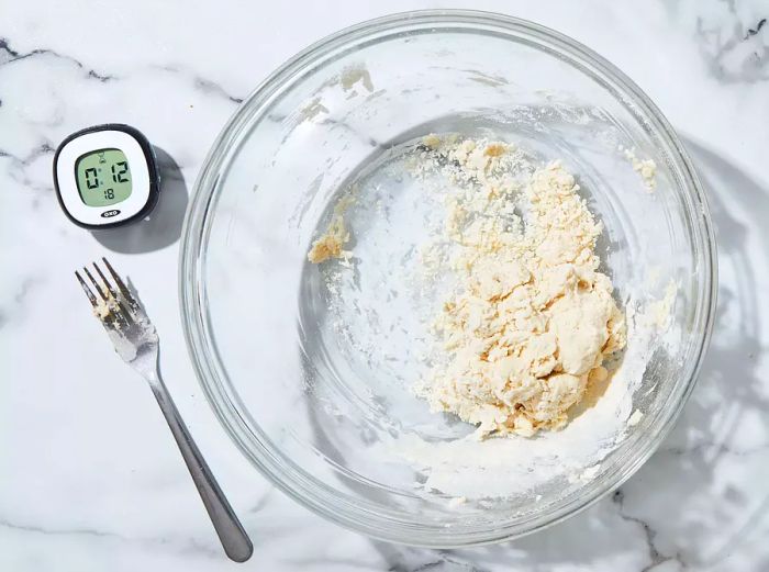 A top-down view of flour and water being mixed together in a bowl, with a fork and timer placed beside it.