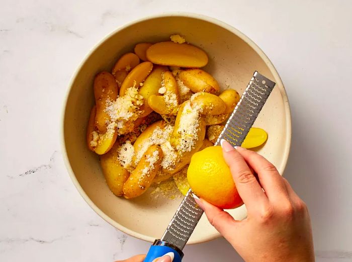 Grating lemon zest over fingerling potatoes with Parmesan cheese in a mixing bowl.