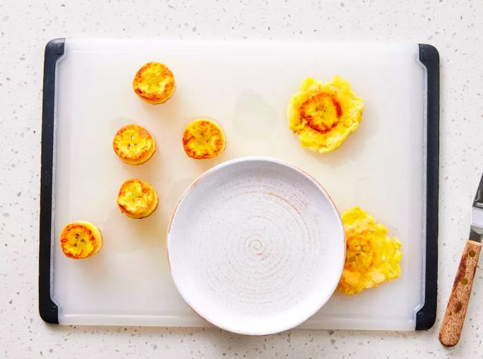 A cutting board with browned plantain slices, one of which is being pressed flat with a plate.