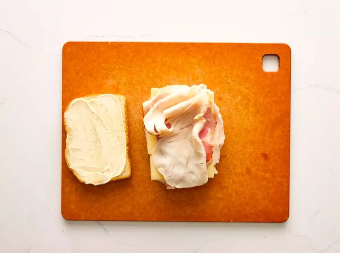 A slice of bread with mustard, topped with layers of ham, turkey, and Swiss cheese.