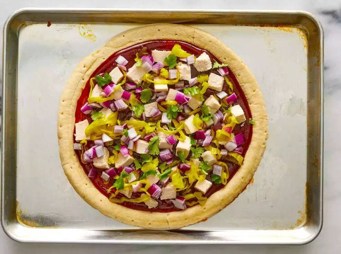 A baking sheet featuring a pizza crust, generously topped with barbecue sauce, chicken, pepperoncini peppers, onions, and cilantro.