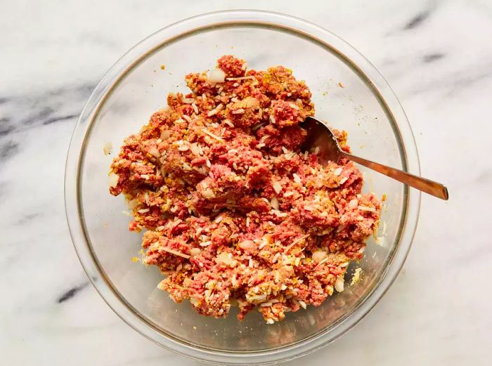 A sizable glass bowl filled with a mixture of ground beef, cooked rice, breadcrumbs, Parmesan cheese, onion, egg, garlic powder, salt, and pepper.