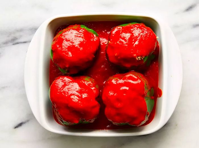 A square baking dish containing four stuffed green bell peppers, all topped with a tangy tomato-vinegar sauce.