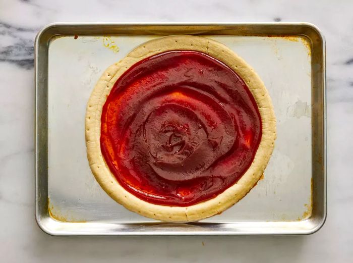 A pizza crust placed on a baking sheet, generously topped with barbecue sauce.