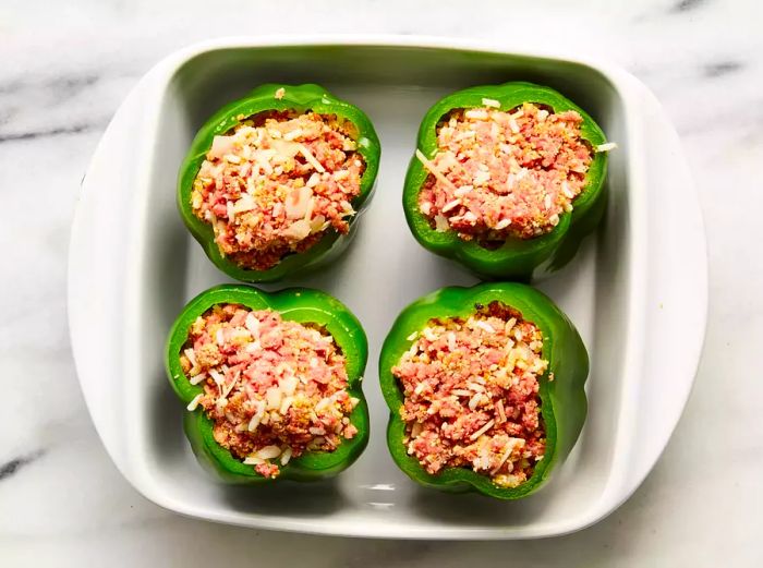 A square baking dish containing four green bell peppers, each stuffed with a savory filling.