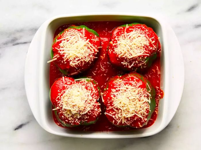 A baking dish with four stuffed green bell peppers, covered in a tomato-vinegar sauce and garnished with Parmesan cheese.