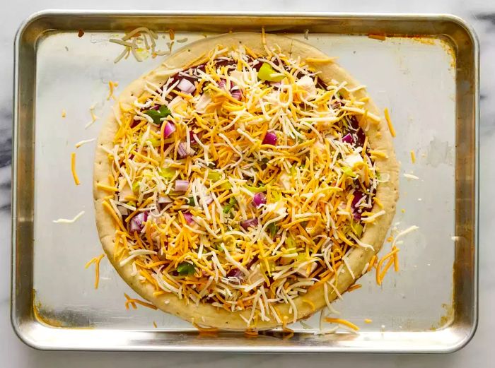 A baking sheet with a pizza crust, covered in barbecue sauce, chicken, pepperoncini peppers, onions, cilantro, and a generous layer of shredded Colby-Jack cheese.