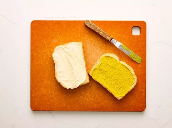 A cutting board displaying one slice of bread with mayonnaise and another with mustard spread on it.