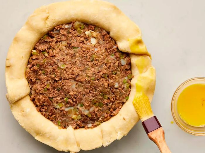 A pie shell filled with meat mixture, with the edges being brushed with egg wash before sealing with the top crust