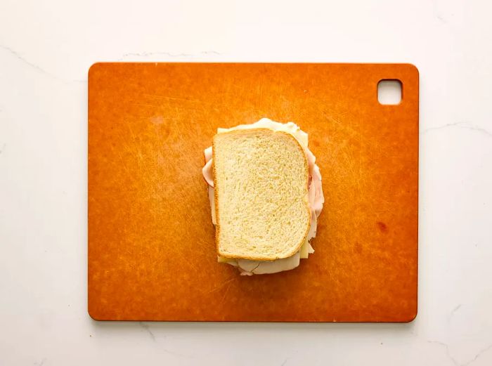 A sandwich with layers of ham, turkey, Swiss cheese, mayonnaise, and mustard resting on a cutting board.