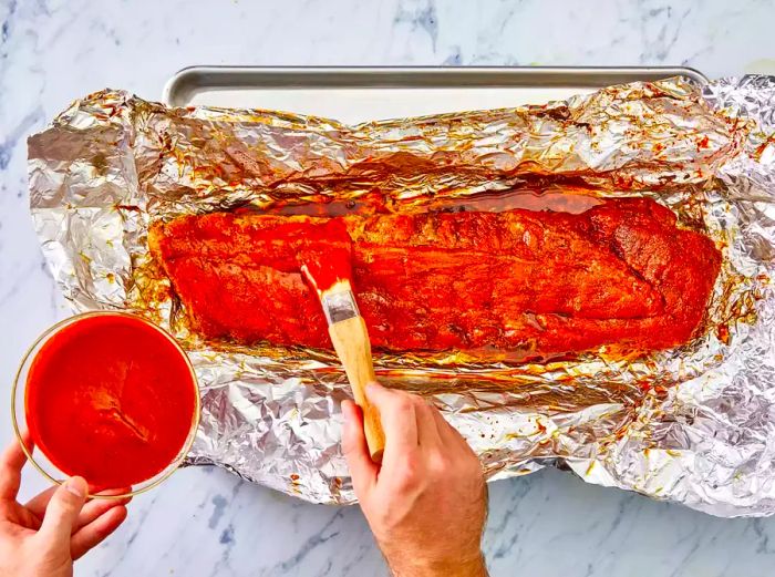 Hands brushing barbecue sauce onto the rack of ribs on a sheet of foil