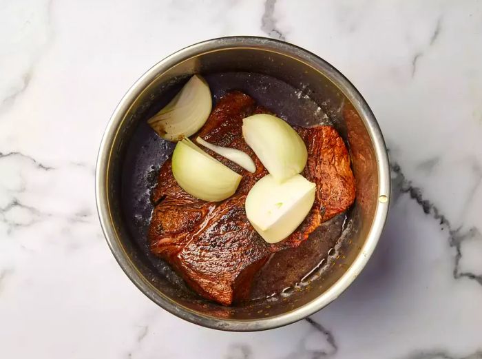 A pressure cooker filled with a seared boneless beef chuck roast, beef broth, quartered onion, and Worcestershire sauce