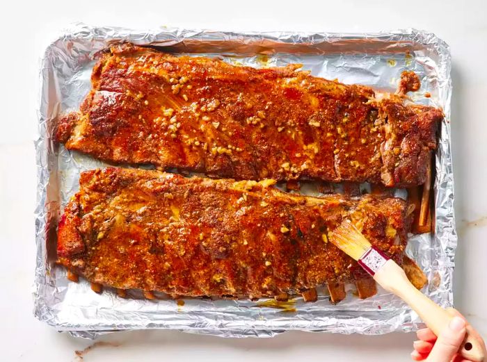 Two racks of pork spareribs being coated with sauce using a brush.