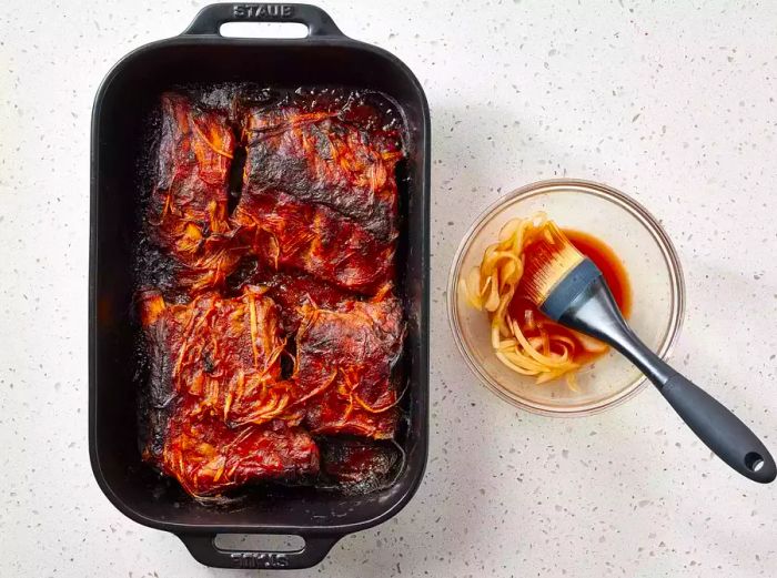 Oven-Baked BBQ Ribs in a roasting pan next to a bowl of sauce and a basting brush