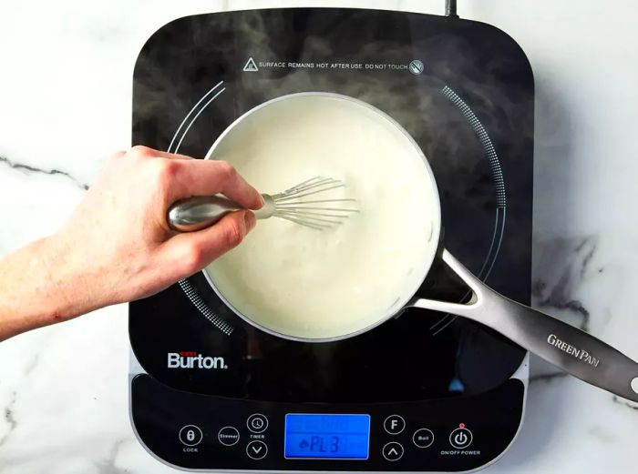 A person whisking milk into the flour-based roux in a small saucepan