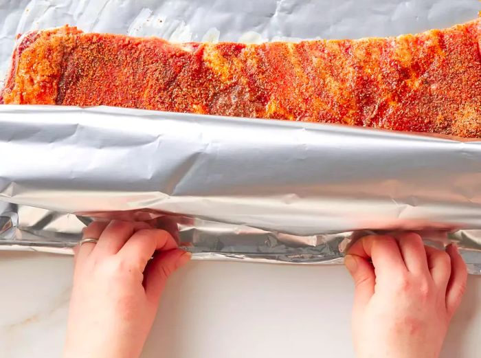 Two hands sealing the pork spare ribs in aluminum foil, ensuring a tight closure.