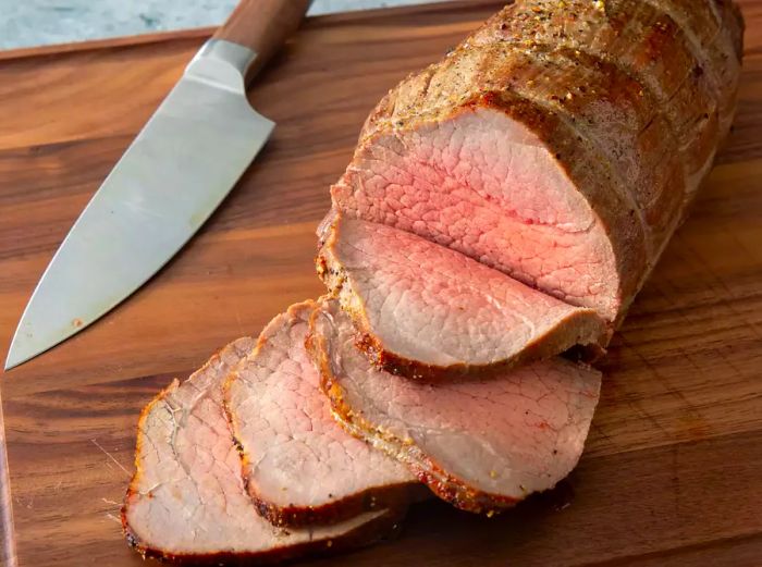 A top-down view of sliced roast beef on a wooden cutting board, next to a knife.