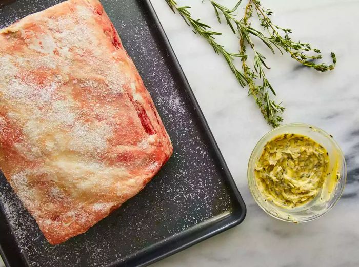 A raw roast resting on a baking sheet, accompanied by a small bowl filled with a mixed herb spread.