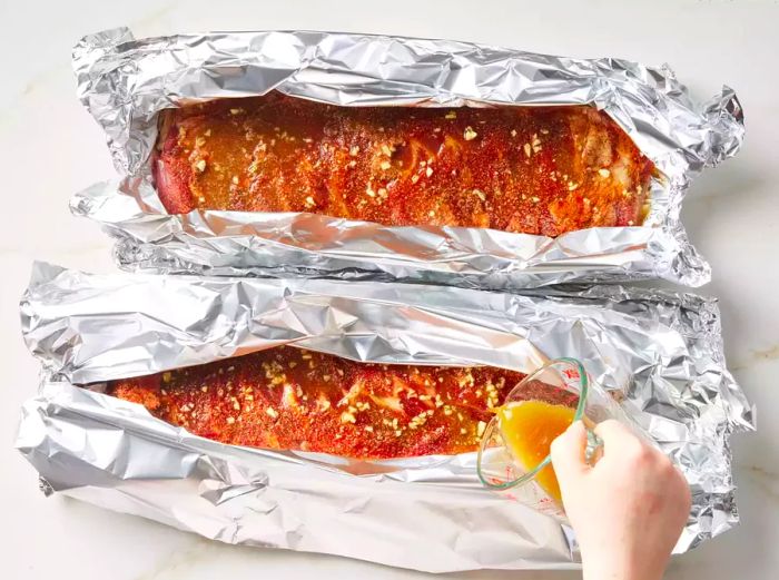 Two partially sealed foil packets containing racks of spareribs, with the beer sauce being poured inside each packet.