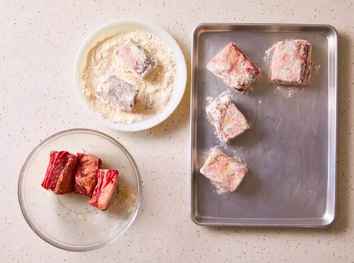 Beef short ribs resting in a bowl, surrounded by a plate of flour and a baking sheet.