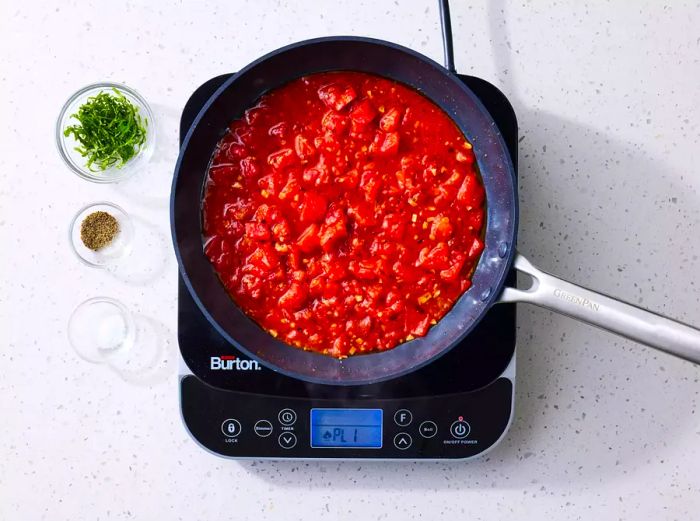 Tomato sauce simmering in a pan on a burner, alongside bowls of shredded basil and seasonings