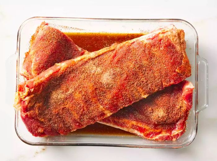 Two racks of seasoned pork spareribs in a glass baking dish, ready to refrigerate.