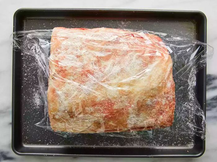 A roast wrapped in plastic wrap, resting on a baking sheet.