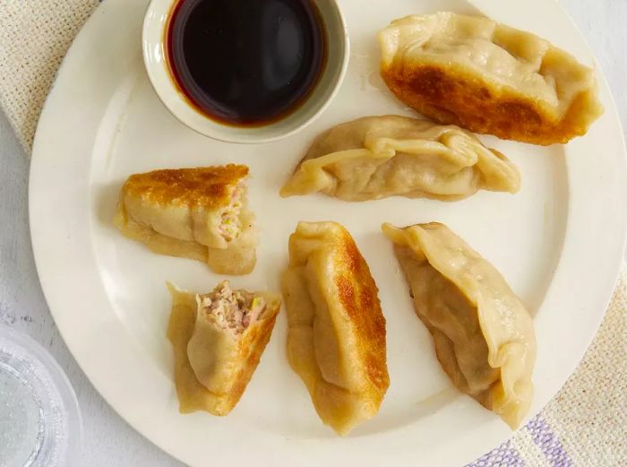 A plate of golden-brown pot stickers, one cut open to reveal the filling, with a side of dipping sauce.