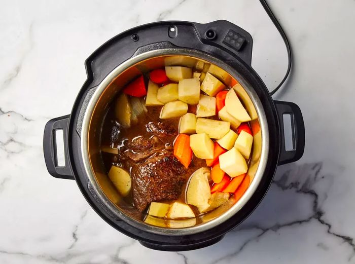 An open pressure cooker filled with beef chuck roast, diced potatoes, and chopped carrots