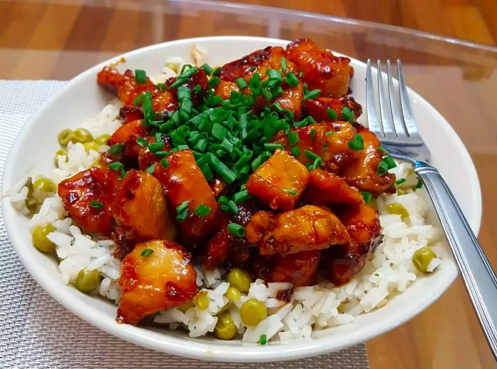 Close-up view of honey-glazed chicken with green onions served on a bed of rice, accompanied by peas