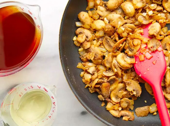 Aerial view of cooked mushrooms and onions in a skillet with cooking wine and broth in measuring cups nearby.