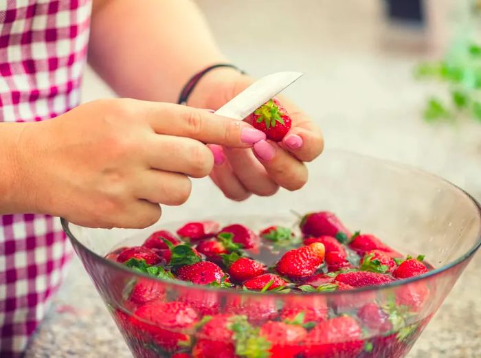 A young woman washes a bowl of fresh strawberries and slices one with a small paring knife.